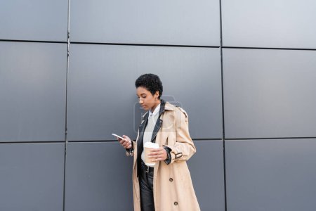 Téléchargez les photos : Jeune femme d'affaires afro-américaine en trench coat beige message sur smartphone et tenant tasse en papier près du mur gris sur la rue - en image libre de droit