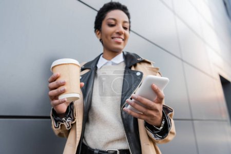 Téléchargez les photos : Femme d'affaires afro-américaine floue avec du café pour aller sourire et des messages sur le téléphone portable près du mur gris sur la rue urbaine - en image libre de droit