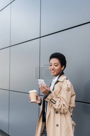 Téléchargez les photos : Joyeuse femme d'affaires afro-américaine en trench coat beige tenant du café dans une tasse en papier et bavardant sur smartphone près d'un mur gris en ville - en image libre de droit