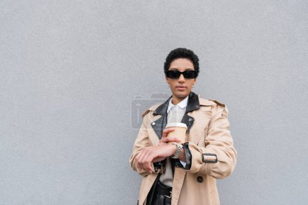 african american businesswoman in sunglasses holding paper cup and checking time on wristwatch while waiting on street