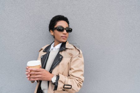 Photo for African american businesswoman in beige trench coat and sunglasses holding paper cup and looking away near grey wall in city - Royalty Free Image