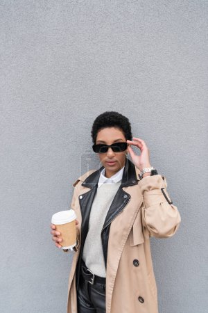 fashionable african american businesswoman in trench coat adjusting sunglasses and holding takeaway drink near grey wall on urban street