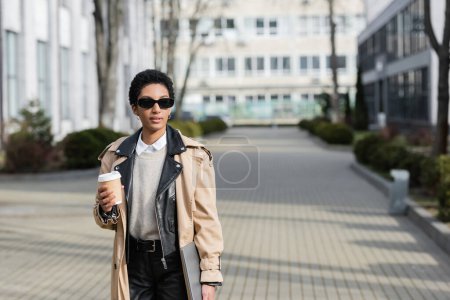 femme d'affaires afro-américaine en lunettes de soleil et élégant trench coat tenant ordinateur portable et café pour aller sur la rue urbaine floue