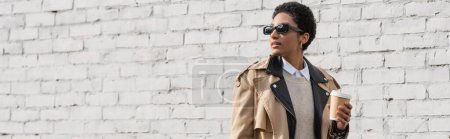 Photo for African american businesswoman in sunglasses and stylish trench coat holding coffee to go and looking away near brick wall on street, banner - Royalty Free Image