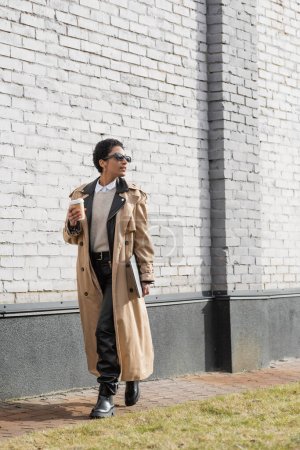 full length of african american businesswoman in sunglasses and trench coat walking with laptop and coffee to go near grey wall on urban street