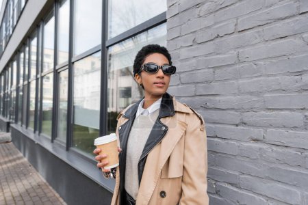 afrikanisch-amerikanische Geschäftsfrau in Sonnenbrille und beigem Trenchcoat hält Imbissgetränk in der Hand und schaut in der Nähe eines Gebäudes an der Stadtstraße weg