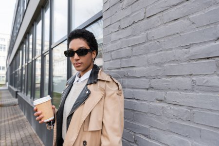 afrikanisch-amerikanische Geschäftsfrau in Sonnenbrille und beigem Trenchcoat steht mit Kaffee in der Nähe einer grauen Hauswand auf der Straße