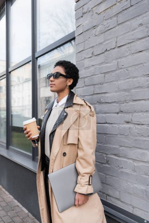 african american businesswoman in stylish trench coat and sunglasses standing with coffee to go and laptop near grey wall of city building 