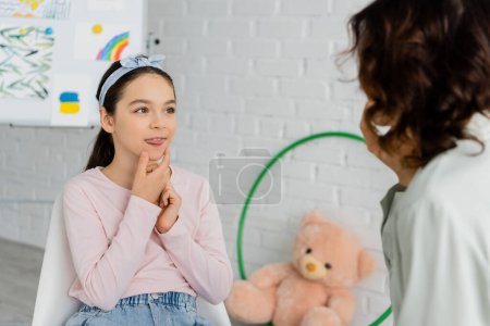 Preteen kid sticking out tongue during lesson with speech therapist in consulting room 