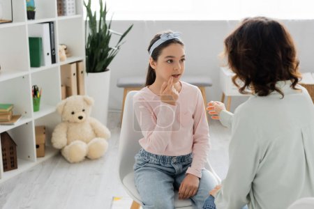 Preteen girl talking and pointing at mouth near blurred speech therapist in consulting room 