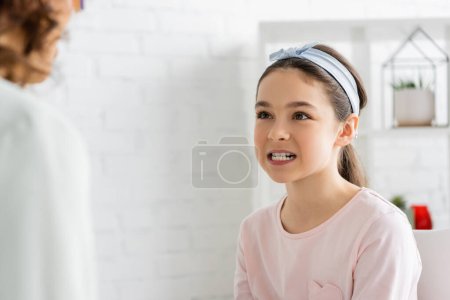 Preteen girl showing teeth near blurred speech therapist in consulting room 