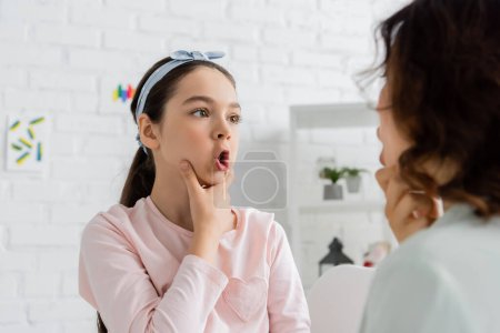 Preteen pupil touching cheeks and talking during lesson with speech therapist 