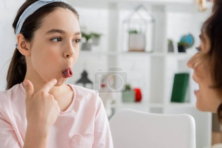Photo for Preteen kid pointing at tongue near blurred speech therapist in consulting room - Royalty Free Image