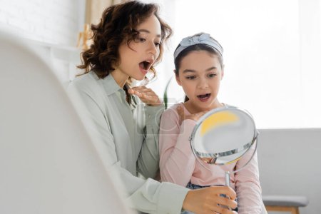 Speech therapist opening mouth near pupil and holding mirror in consulting room 