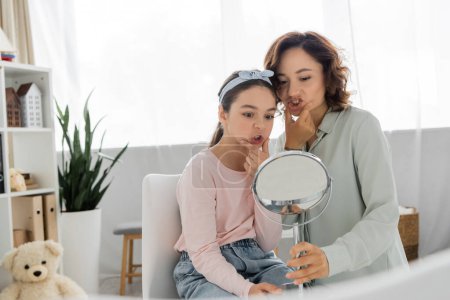Pupil touching cheeks and looking at mirror near speech therapist in consulting room 