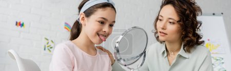 Preteen girl sticking out tongue near mirror and speech therapist in consulting room, banner 