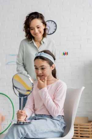 Preteen child holding mirror and speaking near smiling speech therapist in consulting room 