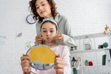Speech therapist touching cheeks of pupil with blurred mirror in consulting room 