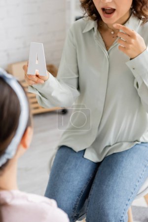 Cropped view of speech therapist holding letter a and pointing at mouth near blurred pupil in consulting room 