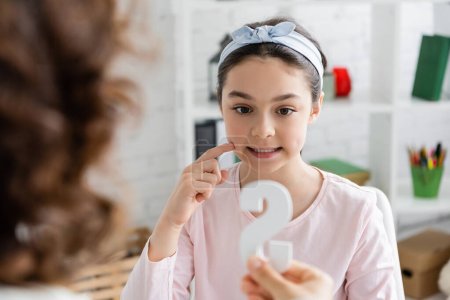 Preteen pupil touching cheek and holding blurred letter s near speech therapist in consulting room 