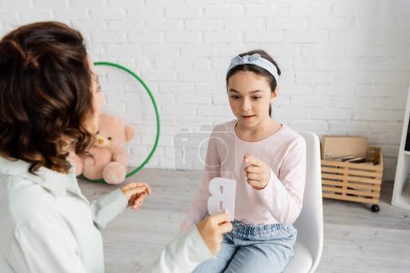 Girl pointing at letter b near blurred speech therapist during lesson in consulting room 