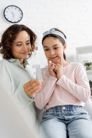 Preteen girl speaking near smiling speech therapist with letter in consulting room 
