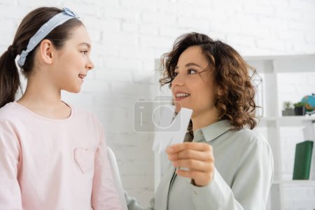 Sonriente terapeuta del habla sosteniendo carta borrosa durante la lección con el niño en la sala de consulta 