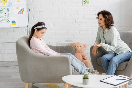 Psychologist talking to sad preteen girl on armchair in consulting room 