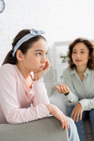 Pensive preteen child sitting near blurred psychologist in consulting room 