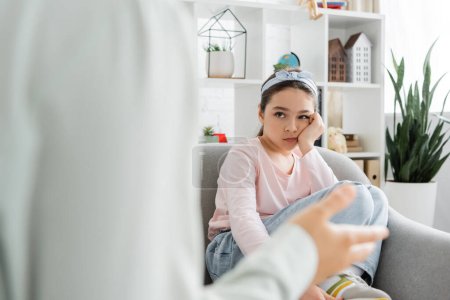 Bored preteen child looking at blurred psychologist in consulting room 