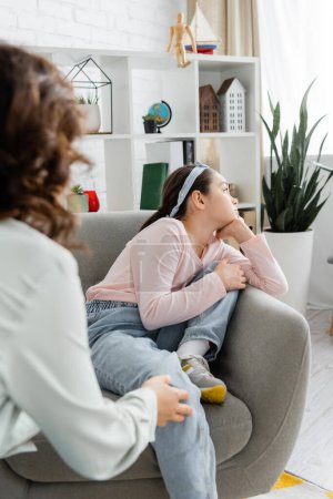 Thoughtful preteen girl looking away while sitting near blurred psychologist in consulting room 