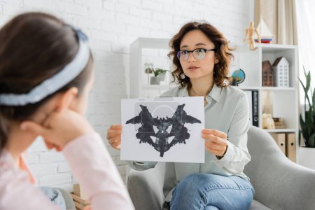Psychologist in eyeglasses holding Rorschach test near blurred child in consulting room 