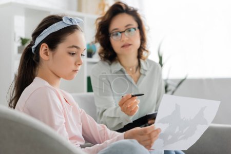 Preteen girl holding Rorschach test near blurred psychologist in consulting room 