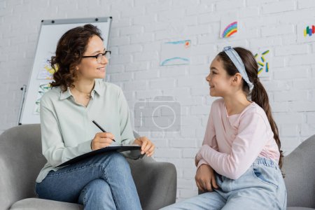 Photo for Smiling psychologist in eyeglasses holding clipboard and looking at preteen daughter in consulting room - Royalty Free Image