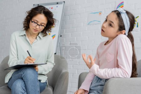 Psychologist writing on clipboard while blurred child talking in consulting room 