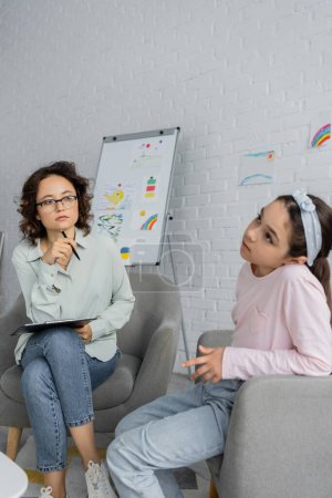 Focused psychologist holding clipboard and looking at pensive child in consulting room 