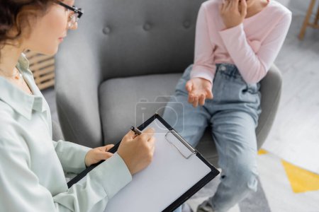 Psychologist writing on clipboard near blurred preteen patient in consulting room 