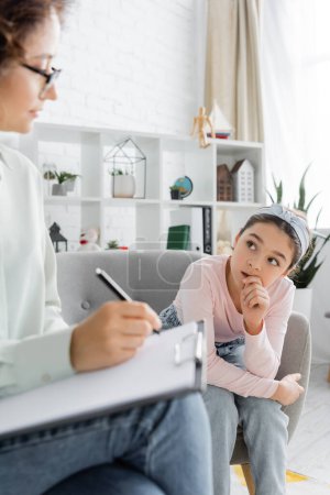 Tensed preteen girl looking at blurred psychologist writing on clipboard in consulting room 