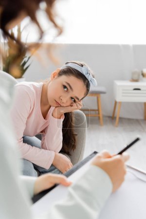 Sad preteen child looking at blurred psychologist with clipboard in consulting room 