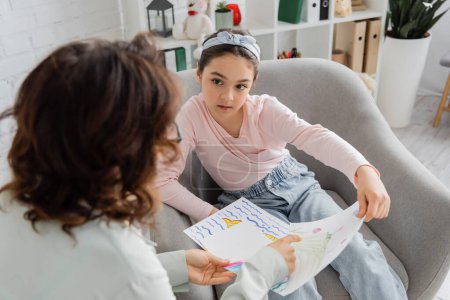 Preteen patient talking to blurred psychologist pointing at drawing in consulting room 