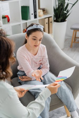 Preteen girl talking and pointing at drawings near blurred psychologist in consulting room 