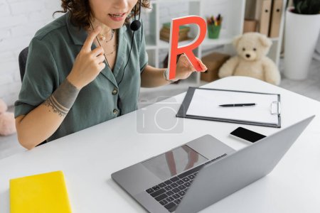 Cropped view of speech therapist in headset pointing at mouth and holding letter during online lesson 