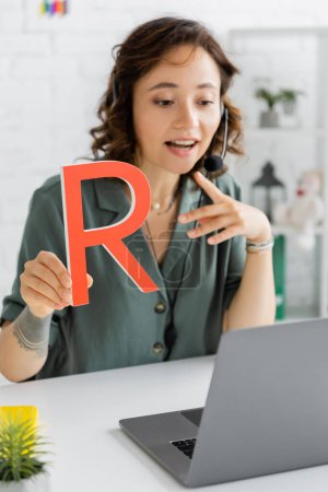 Blurred speech therapist in headset holding letter during online lesson in consulting room 