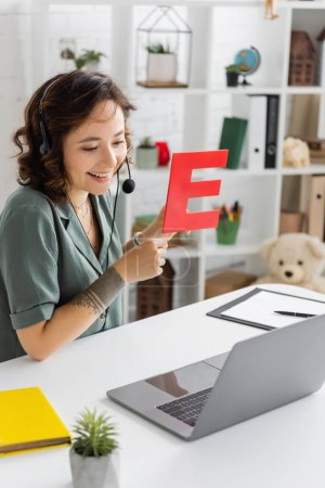 Überglücklicher Logopäde im Headset hält Brief während Videoanruf auf Laptop im Sprechzimmer 
