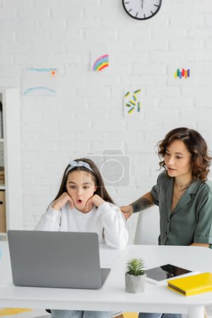 Girl having speech therapy video call on laptop near mother at home 