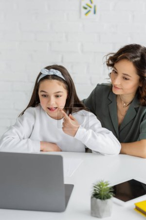 Preteen girl pointing at mouth during speech therapy online lesson near mom at home 