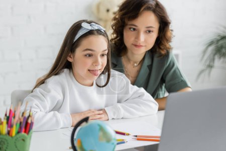 Preteen Mädchen streckt Zunge während Logopädie Online-Lektion in der Nähe von Mama zu Hause 