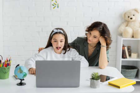 Preteen girl sticking out tongue during speech therapy online lesson on laptop near mother at home 