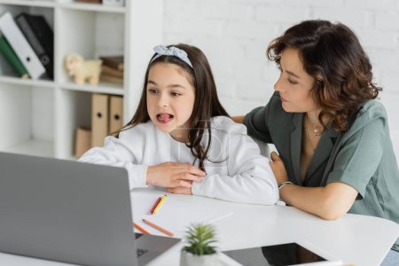 Parent sitting near daughter having speech therapy online lesson near devices at home 