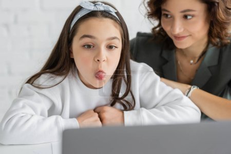 Preteen girl sticking out tongue during speech therapy lesson on laptop near blurred mom at home 